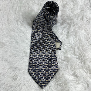 Puede incluir: Una corbata azul oscuro con un patrón repetitivo de motivos de estribos ecuestres en dorado y blanco. La corbata está doblada en la parte superior, mostrando un extremo puntiagudo y una etiqueta de tela visible en el reverso de la pala.
