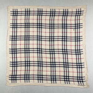 May include: Square scarf in cream with a navy, red, and black plaid pattern. The scarf has a border and a subtle sheen. The Burberry logo is visible in the bottom right corner.