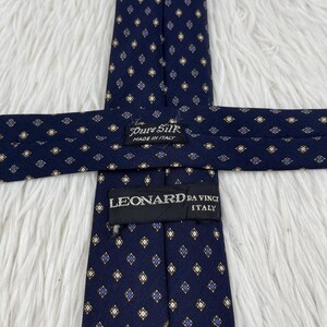K&ouml;nnte beinhalten: Eine marineblaue Krawatte mit einem sich wiederholenden Muster aus kleinen goldenen und blauen Rauten. Die Krawatte hat ein schwarzes Etikett mit der Aufschrift Pure Silk Made in Italy und ein zweites schwarzes Etikett mit LEONARD DA VINCI ITALY.