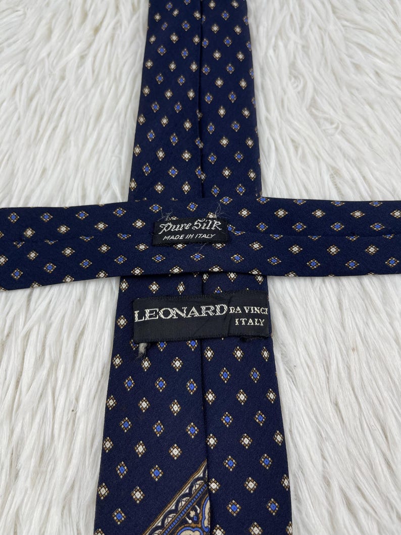 K&ouml;nnte beinhalten: Eine marineblaue Krawatte mit einem sich wiederholenden Muster aus kleinen goldenen und blauen Rauten. Die Krawatte hat ein schwarzes Etikett mit der Aufschrift Pure Silk Made in Italy und ein zweites schwarzes Etikett mit LEONARD DA VINCI ITALY.