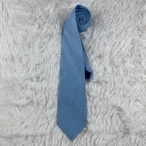 May include: A pale blue tie with a small, dotted design. The tie is rolled at the top and rests on a white, fluffy background. The tie's colour is a soft, muted blue, and the fabric has a slight sheen.