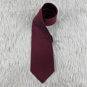 May include: A necktie featuring a deep burgundy red base color with thin, evenly spaced diagonal black stripes. The tie is folded at the top, showing the inner lining and the consistent striped pattern across the entire length of the fabric.