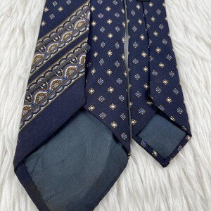 K&ouml;nnte beinhalten: Eine marineblaue Krawatte mit einem sich wiederholenden Muster aus kleinen goldenen und blauen Rauten. Eine Seite zeigt ein dekoratives diagonales Band mit komplizierten, vom Paisley-Muster inspirierten Details in Gold, Blau und Wei&szlig;. Ein schwarzes Etikett tr&auml;gt die Aufschrift Pure Silk Made in Italy.