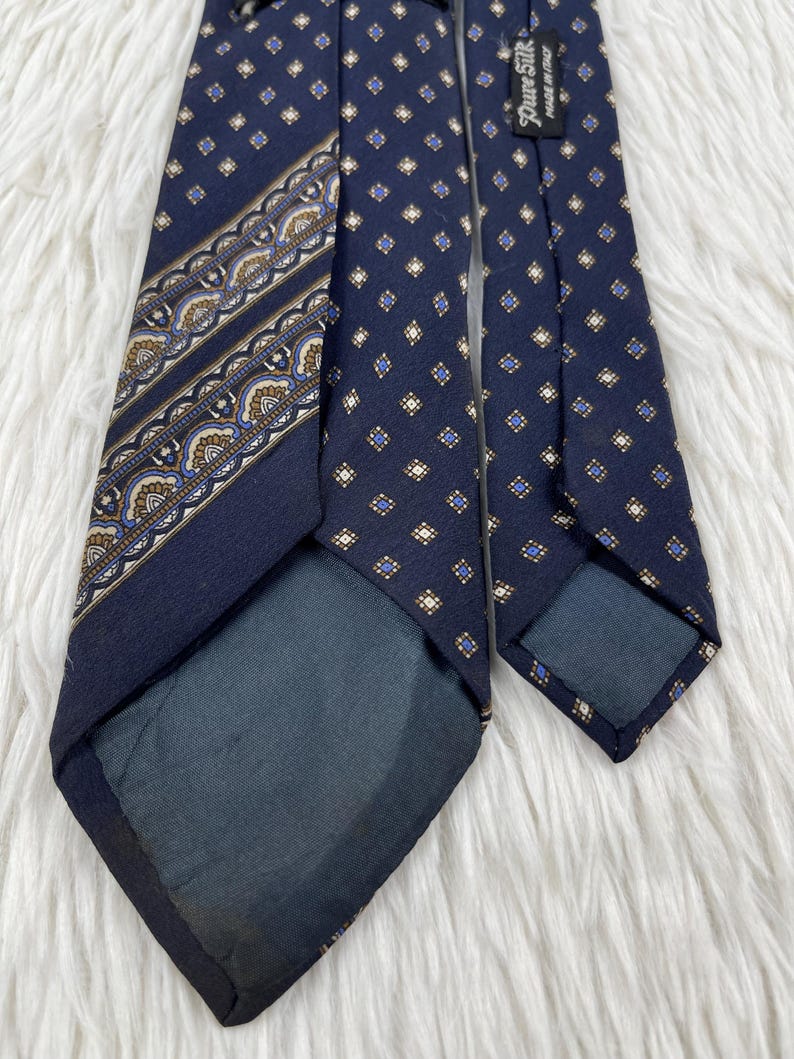 K&ouml;nnte beinhalten: Eine marineblaue Krawatte mit einem sich wiederholenden Muster aus kleinen goldenen und blauen Rauten. Eine Seite zeigt ein dekoratives diagonales Band mit komplizierten, vom Paisley-Muster inspirierten Details in Gold, Blau und Wei&szlig;. Ein schwarzes Etikett tr&auml;gt die Aufschrift Pure Silk Made in Italy.