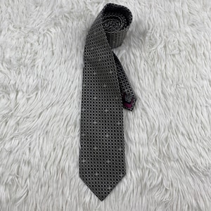 Puede incluir: Una corbata gris oscuro con un patrón geométrico de pequeños puntos blancos. La corbata está enrollada en la parte superior y descansa sobre una superficie blanca texturizada. El extremo de la corbata tiene una etiqueta rosa.