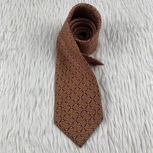 May include: A necktie featuring a deep burgundy background with a repeating gold and white geometric Celtic knot pattern. The tie is rolled at the top, showing the fabric texture and the intricate, interconnected line design across the entire surface.