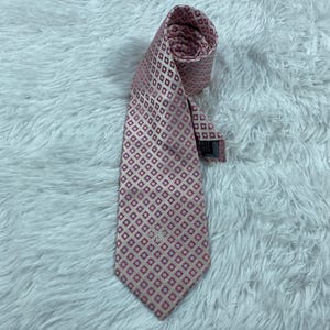 May include: A rolled-up pink tie with a repeating geometric pattern of small red squares on a light pink background. The tie is made of a shiny material and has a label at the end.