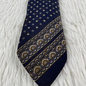 K&ouml;nnte beinhalten: Eine dunkelblaue Krawatte mit einem sich wiederholenden Muster aus kleinen hellen Quadraten im oberen Bereich und einem breiten diagonalen Streifen im unteren Bereich, der mit einem ornamentalen Bogenmotiv in Beige, Blau und Wei&szlig; verziert ist.