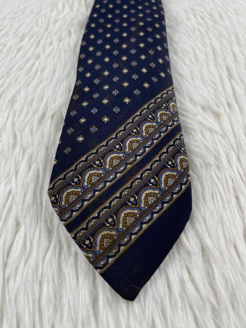 K&ouml;nnte beinhalten: Eine dunkelblaue Krawatte mit einem sich wiederholenden Muster aus kleinen hellen Quadraten im oberen Bereich und einem breiten diagonalen Streifen im unteren Bereich, der mit einem ornamentalen Bogenmotiv in Beige, Blau und Wei&szlig; verziert ist.