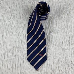 May include: A navy blue necktie featuring a repeating pattern of diagonal cream-colored stripes that resemble film strips. The lower portion of the tie displays a small, centered red and green emblem with a gold-toned logo detail.