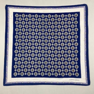 Pode incluir: Um lenço quadrado azul marinho com um padrão geométrico repetitivo em castanho, branco e verde. Tem uma borda branca com acabamento roxo e azul marinho. O texto "ALFREDO VERSACE" está impresso em branco.