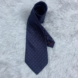 May include: A navy blue necktie featuring a subtle herringbone weave pattern and a repeating grid of small, light-colored square dots. The tie is rolled at the top and displays a pointed tip at the bottom, with a dark fabric label visible on the back.
