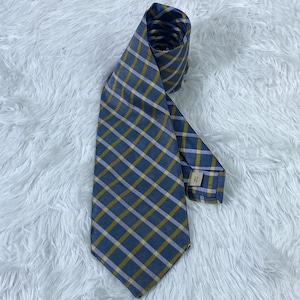 May include: A rolled-up necktie with a plaid pattern in shades of blue, yellow, and lavender. The tie is made of a smooth fabric and is displayed on a white, fluffy surface. The tie's design features a classic plaid pattern.