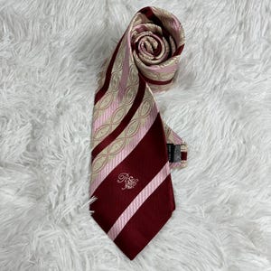 May include: A necktie featuring a diagonal striped pattern in deep burgundy, light pink, and beige. The beige sections display an intricate oval geometric design. The lower portion of the tie includes an embroidered monogram logo in light pink thread.
