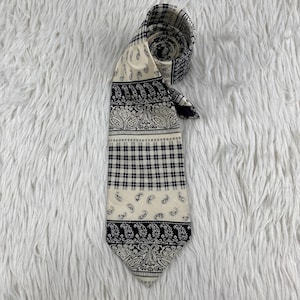 May include: A necktie featuring a cream-coloured background with a black printed pattern. The design consists of horizontal bands alternating between intricate paisley motifs, a small square grid pattern, and scattered paisley elements throughout.