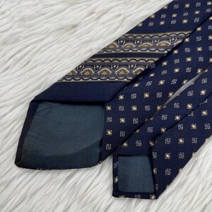 K&ouml;nnte beinhalten: Eine marineblaue Krawatte mit einem sich wiederholenden Muster aus kleinen wei&szlig;en und blauen Quadraten, akzentuiert durch ein horizontales Band mit einem dekorativen geometrischen Design in Braun, Wei&szlig; und Blau. Die Krawatte hat ein dunkelblaues Futter.