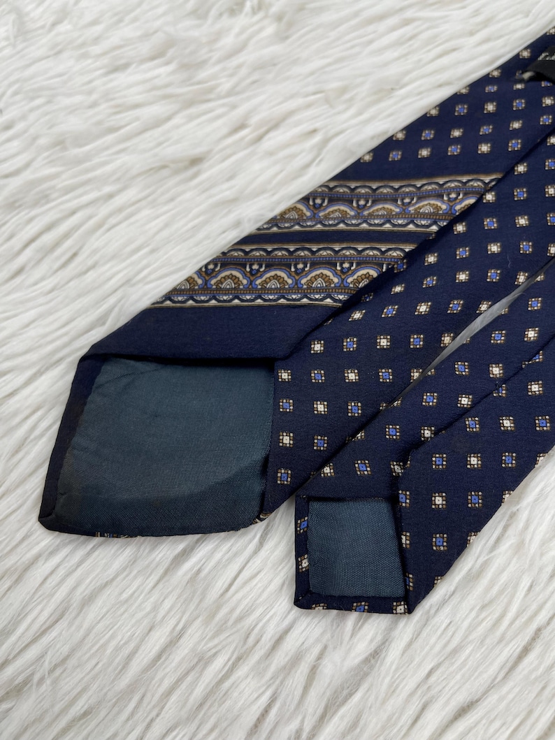 K&ouml;nnte beinhalten: Eine marineblaue Krawatte mit einem sich wiederholenden Muster aus kleinen wei&szlig;en und blauen Quadraten, akzentuiert durch ein horizontales Band mit einem dekorativen geometrischen Design in Braun, Wei&szlig; und Blau. Die Krawatte hat ein dunkelblaues Futter.