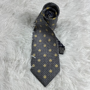 May include: A necktie featuring a dark navy blue background with a repeating grid pattern of small white squares. Within the grid, there are evenly spaced, small golden yellow house-shaped icons. The tie is rolled at the top and displays a label.
