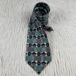 Peut inclure: Une cravate présentant un motif géométrique répétitif composé d'ovales, de triangles et de carrés bleu foncé sur des tons vert sauge, bleu clair et taupe. La cravate est enroulée sur le haut, révélant une structure en tissu avec une extrémité pointue.