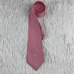 May include: A pink necktie featuring a subtle pattern of tiny white dots and a small embroidered logo near the pointed tip. The fabric has a fine diagonal weave texture, and the top portion of the tie is neatly rolled into a circular shape.