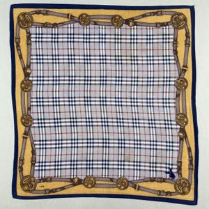 May include: Square scarf with a plaid pattern in blue, white, and red. The scarf has a gold border with a design of belts and medallions. A dark blue border frames the scarf.