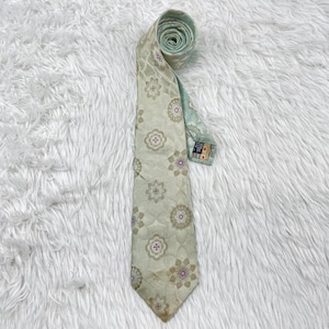 Puede incluir: Una corbata vintage con un estampado floral en tonos beige, verde y morado. La corbata está enrollada en la parte superior, revelando un forro verde claro. La tela tiene un brillo sutil.