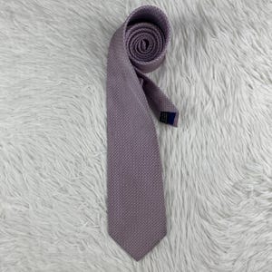 May include: A lavender necktie featuring a subtle, repeating geometric micro-pattern across the entire surface. The tie is rolled at the top, revealing a dark blue fabric label stitched onto the inner lining near the neck area of the garment.