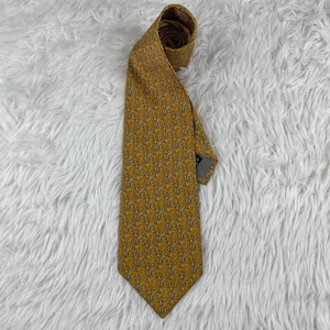 May include: Mustard yellow tie with a repeating pattern of stylized figures in a darker shade. The tie is made of a smooth fabric and is knotted at the top, with the pointed end visible. The background is a white, fluffy surface.