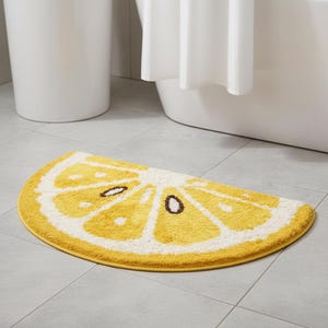 May include: A yellow and white bath mat shaped like a lemon slice. The mat has a yellow border and a detailed design of a lemon's segments, with brown seed details. The mat is placed on a tiled floor near a bathtub.
