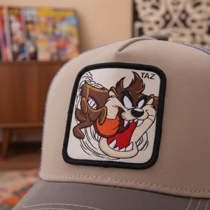 May include: A tan and gray trucker hat featuring a patch with the cartoon character Taz. The patch has a black border and depicts Taz with a red mouth, brown fur, and the word "TAZ" at the top. The hat has a mesh back.