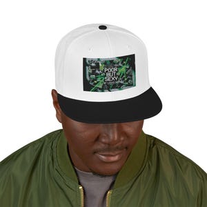 Op de afbeelding: Een wit en zwarte snapback pet met de tekst "POOR BUT SEXY" in een groen en zwarte afbeelding. De pet heeft een zwarte klep en een witte kroon. De pet wordt gedragen door een persoon die een groene jas draagt.