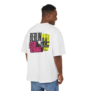 May include: White t-shirt with a graphic on the back. The graphic includes the text "BERLIN STAY STRANGE" in black and pink, and an image of the Berlin TV Tower. The shirt has short sleeves and a loose fit.
