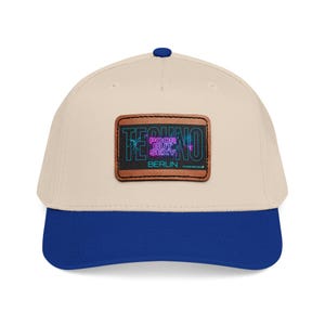 Puede incluir: Gorra de béisbol con corona color crema y visera azul real. La gorra presenta un parche de cuero marrón con las palabras "TECHNO POOR BUT SEXY BERLIN" en una fuente de estilo neón.