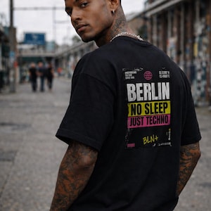 May include: Black t-shirt with the text "BERLIN NO SLEEP JUST TECHNO" in white, yellow, and pink. The shirt also features the text "BLN+" and coordinates. The model is wearing a silver chain and bracelet.