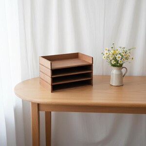 May include: A wooden desk organizer with four stacked trays. The organizer is made of light brown wood and sits on a wooden table. A small vase with white and yellow flowers is to the right of the organizer.