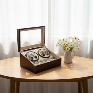 May include: A brown wooden watch winder with a glass top, showcasing four watches on rotating stands. The winder is on a wooden table, with a vase of white flowers in the background. The winder has a beige interior.