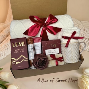 Könnte beinhalten: Eine Geschenkbox mit einer weißen Strickdecke mit dunkelroter Satinschleife, einer Packung Lumi cremiger heißer Schokolade, veganer Handcreme, einem Glasröhrchen mit Badesalz, einer dunkelroten Kunstrose, Karamellbonbons und einer Tasse.