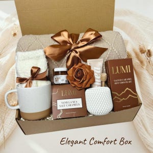 Puede incluir: Una caja de regalo que contiene una manta beige texturizada con un lazo de satén marrón, una taza blanca moteada, calcetines blancos suaves, un tarro de miel, un cucharón de madera, una rosa marrón, un tubo de crema y chocolate caliente.
