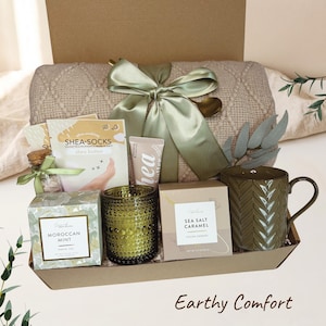 Könnte beinhalten: Eine Geschenkbox mit einer beigen Strickdecke mit Zopfmuster, die mit einem salbeigrünen Band gebunden ist, einem grünen strukturierten Becher, einem grünen Glas-Kerzenhalter, marokkanischem Minztee, Salzkaramell-Keksen, Sheabutter-Creme und Socken.