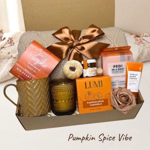 May include: A gift box containing a beige knit blanket with a brown satin bow, a textured brown mug, a glass candle, a small pumpkin, a jar of honey, a box of Spicy Pumpkin tea, Lumi pumpkin spice caramels, Pedi in a Box, and pumpkin pie hand cream.