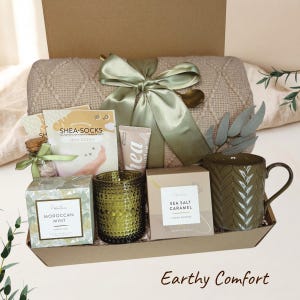 May include: A gift box containing a beige cable-knit blanket tied with a sage green ribbon, a green textured mug, a green glass candle holder, a box of Moroccan Mint tea, a box of Sea Salt Caramel cookies, shea butter cream, and shea socks.