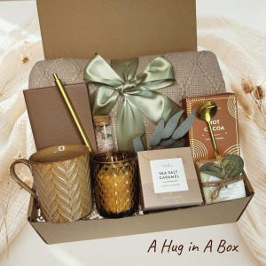 Puede incluir: Una caja de regalo que contiene una manta de punto beige con un lazo verde salvia, un cuaderno marrón, un bolígrafo dorado, un frasco de vidrio, una taza marrón texturizada, una vela de vidrio oscuro, galletas de caramelo y chocolate caliente.