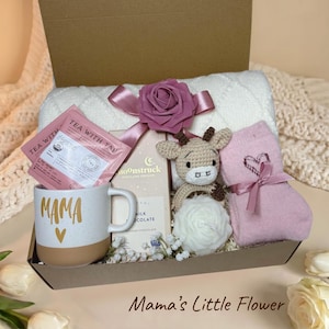 Könnte beinhalten: Eine Geschenkbox mit einer weiß-beigen Tasse mit der Aufschrift MAMA und einem Herz, einem Tea With Tae Teebeutel, einer Moonstruck Milchschokoladentafel, einem gehäkelten Tier, einer weißen blumenförmigen Kerze und rosa Socken mit Herzmuster.