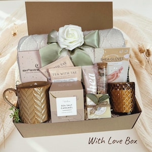 Könnte beinhalten: Eine Geschenkbox mit Pflegeprodukten. Sie enthält eine Tasse, Tee, Sheabutter-Handschuhe, eine Kerze und andere Schönheitsprodukte. Eine weiße Rose und ein grünes Band schmücken die Oberseite der Box. Der Text "With Love Box" steht unten.