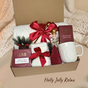 Könnte beinhalten: Eine Geschenkbox mit einer weißen, strukturierten Decke mit roter Satinschleife, einem weißen Becher mit Zopfmuster, einem Nussknacker-Ornament, einer roten Schachtel Lumi Creamy Hot Chocolate und einer Schachtel Lumi Vanille-Meersalz-Karamellen.