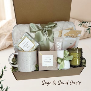 Op de afbeelding: Een geschenkdoos met een witte, getextureerde mok, een doos Moroccan Mint thee, Sea Salt Caramel koekjes, een tube sheaboter, Shea-Socks, een witte handdoek met een saliegroen lint, een groene glazen kaars en een witte deken.
