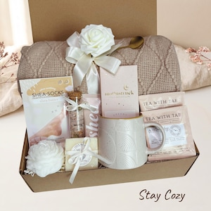 Op de afbeelding: Een geschenkdoos met een beige gebreide deken met een wit rozenlint, een gespikkelde keramische mok, twee Tea With Tae theezakjes, een Moonstruck chocoladereep, een pakje shea butter sokken, een glazen flesje badzout en een witte bloemvormige zeep.