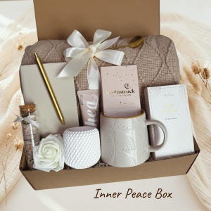 Op de afbeelding: Een cadeaupakket gevuld met zelfzorgartikelen. Inbegrepen zijn een beige deken, een gouden pen, een notitieboek, een mok, een kandelaar, een roos, een flesje met een kurk, een tube lotion en twee dozen met lekkernijen. De doos is vastgebonden met een witte strik. Tekst op de doos luidt "Inner Peace Box."