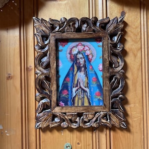 May include: A rectangular wooden frame with intricate carved leaf patterns surrounding a print of a person with a floral crown and a blue head covering. The person is depicted with hands pressed together in a prayer gesture against a blue background.