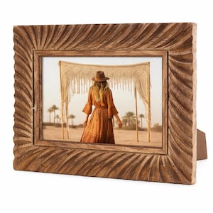 May include: A wooden picture frame with a carved, textured design. The frame holds a photograph of a person in a dress and hat, standing in a desert landscape. The frame has a built-in stand.